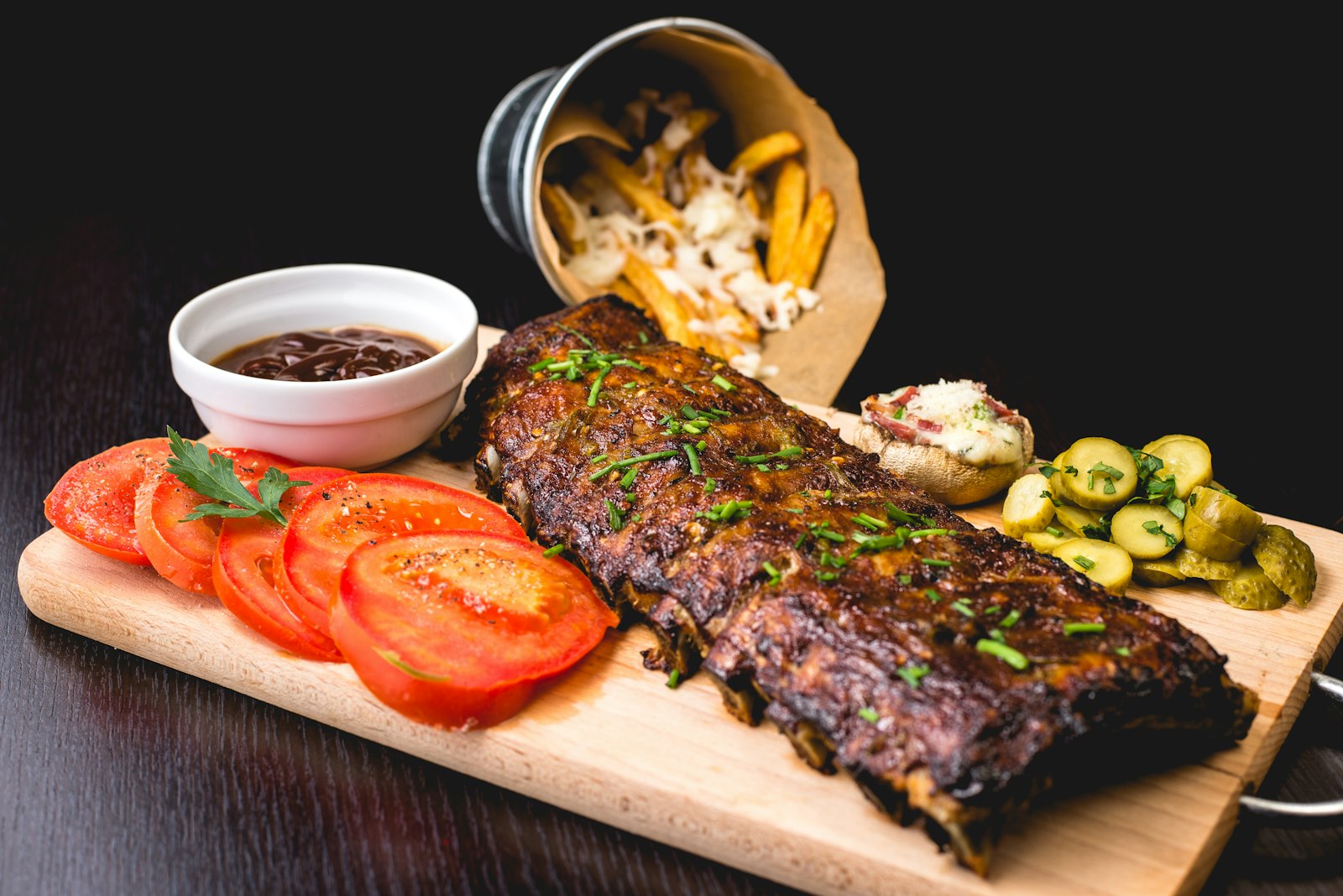 Glazed rack of ribs on a wooden board with sliced tomatoes, fries and dipping sauce