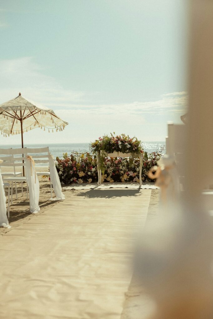 Floral arch and a paper parasol at the end of a sand aisle facing the sea