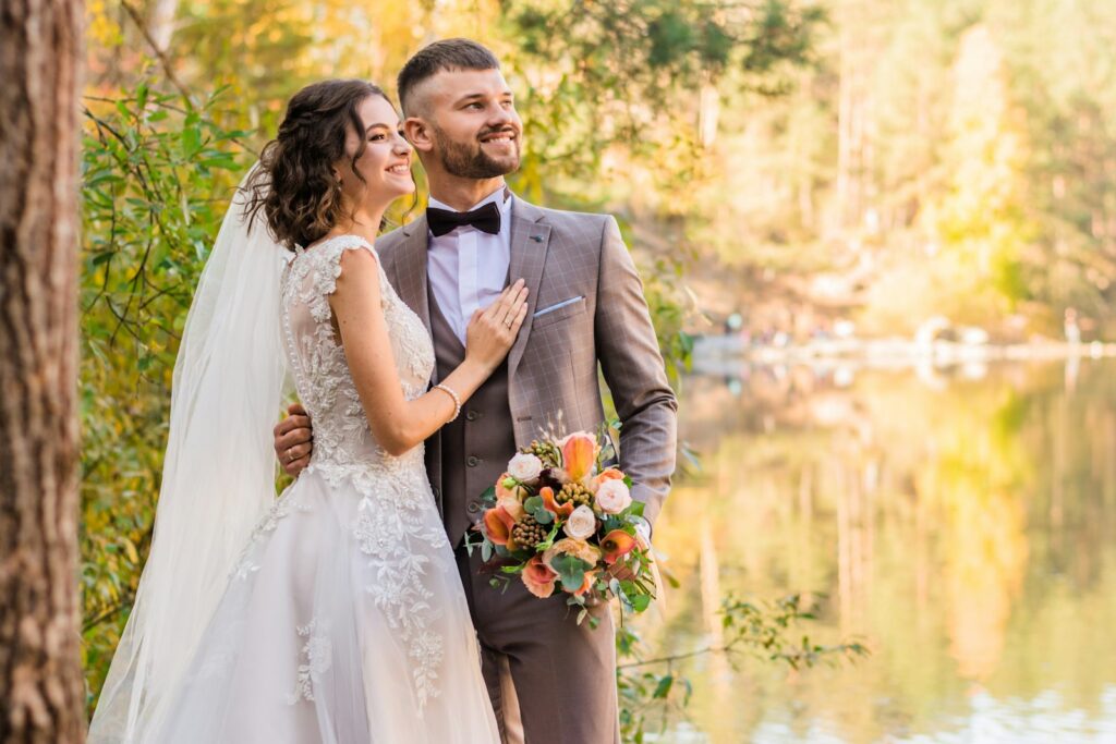 Bride with a peach bouquet leaning on the groom beside an autumn lake