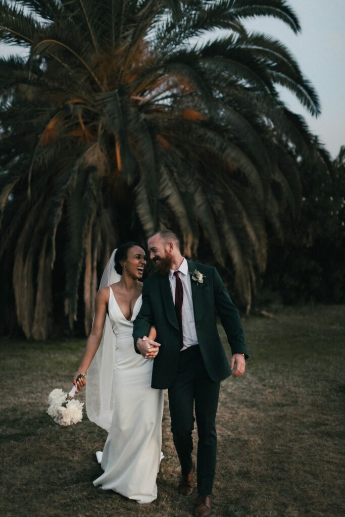 Bride and groom walking across a lawn beneath a palm tree
