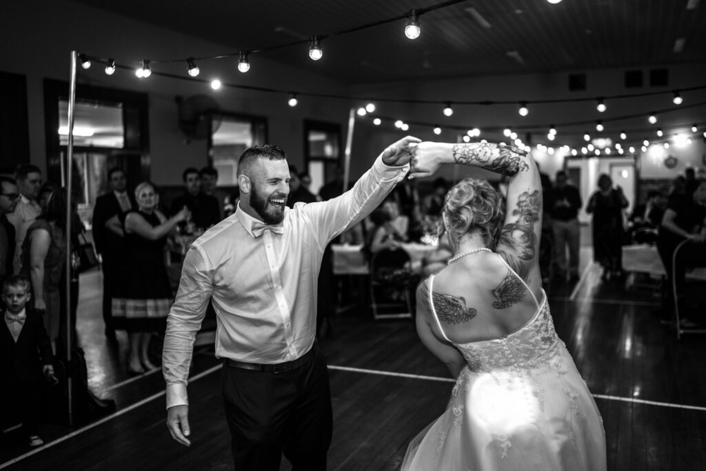 Groom spinning the bride on a dance floor under festoon lights, in monochrome