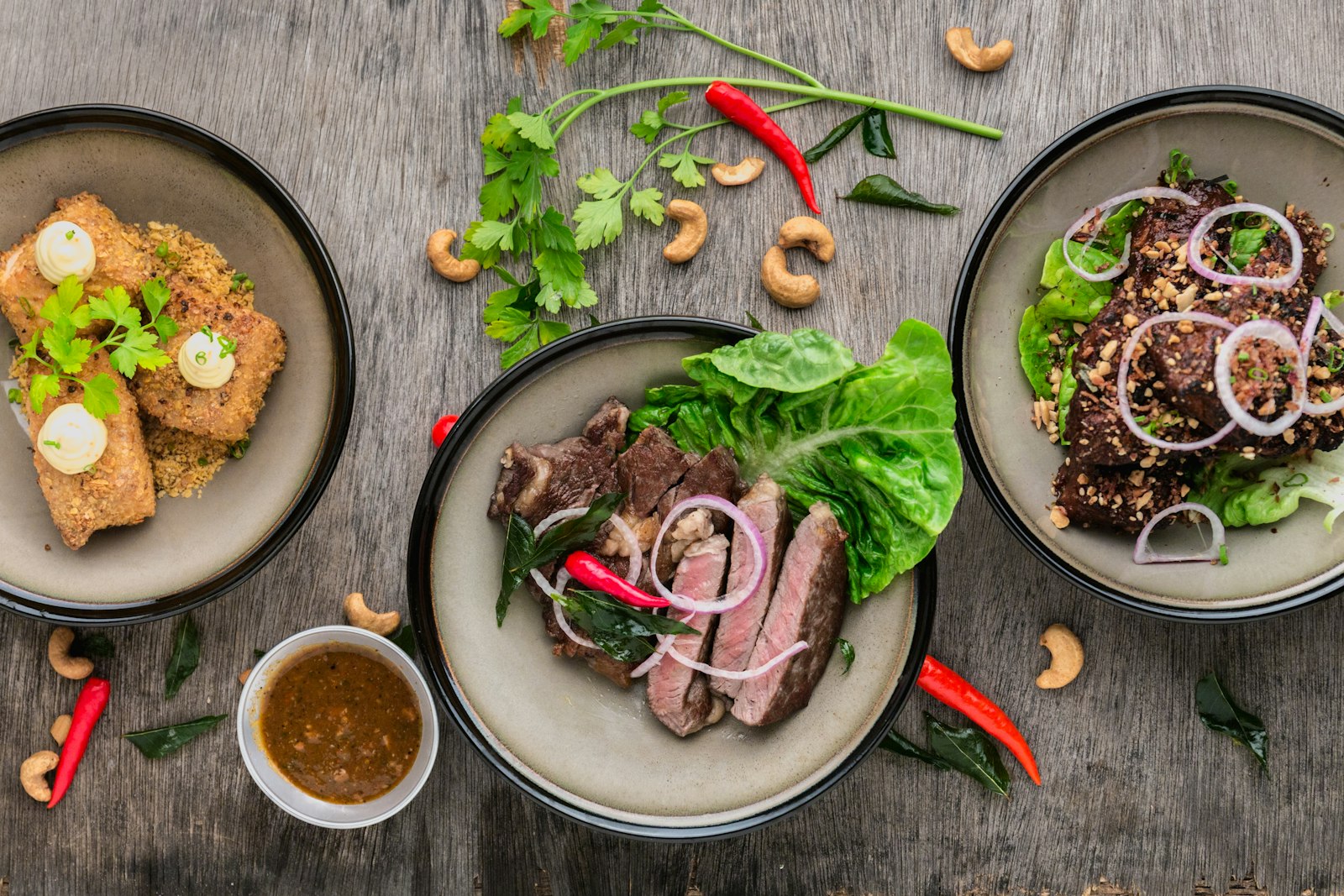 Overhead of three bowls with seared sliced beef, salad and dipping sauce on a dark wooden table