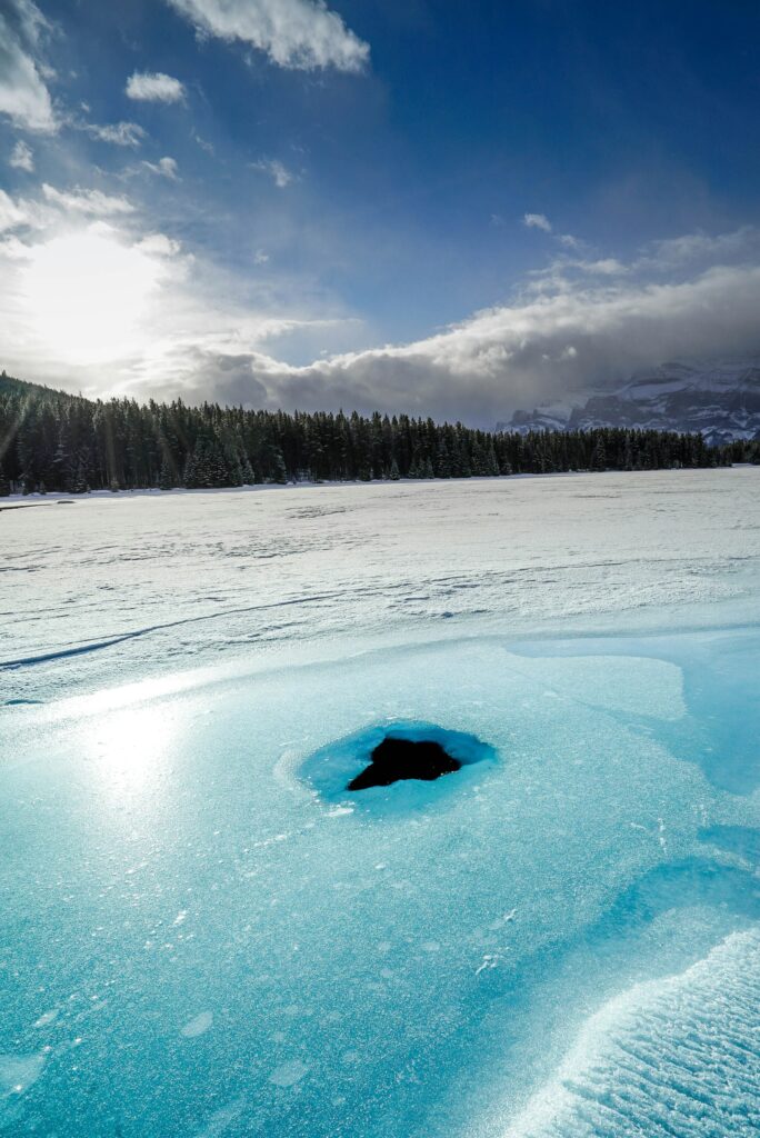 Dark hole in the turquoise ice of a frozen lake, pine forest and mountains beyond