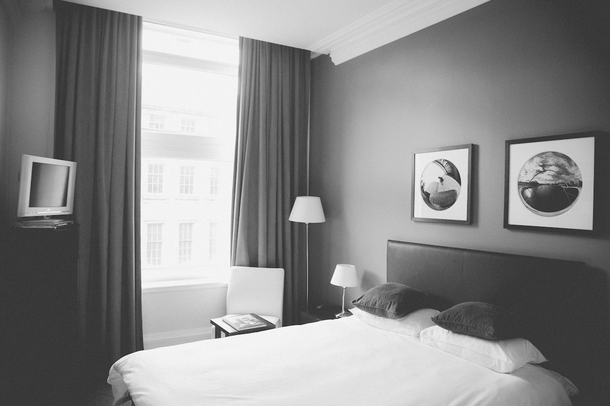 Hotel bed beside a tall window with daylight and a stone building outside