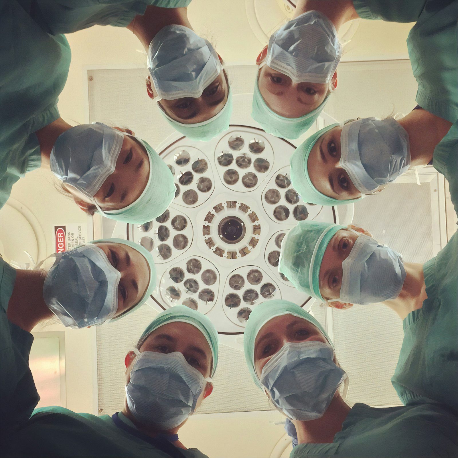 Masked surgical team in a circle looking down at the camera beneath an operating light