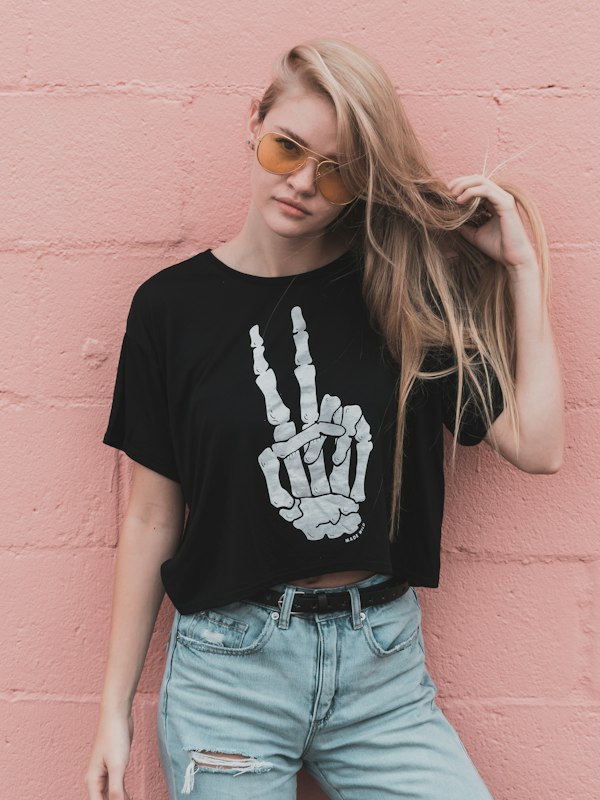Blonde woman in sunglasses and a skeleton-hand tee against a pink brick wall