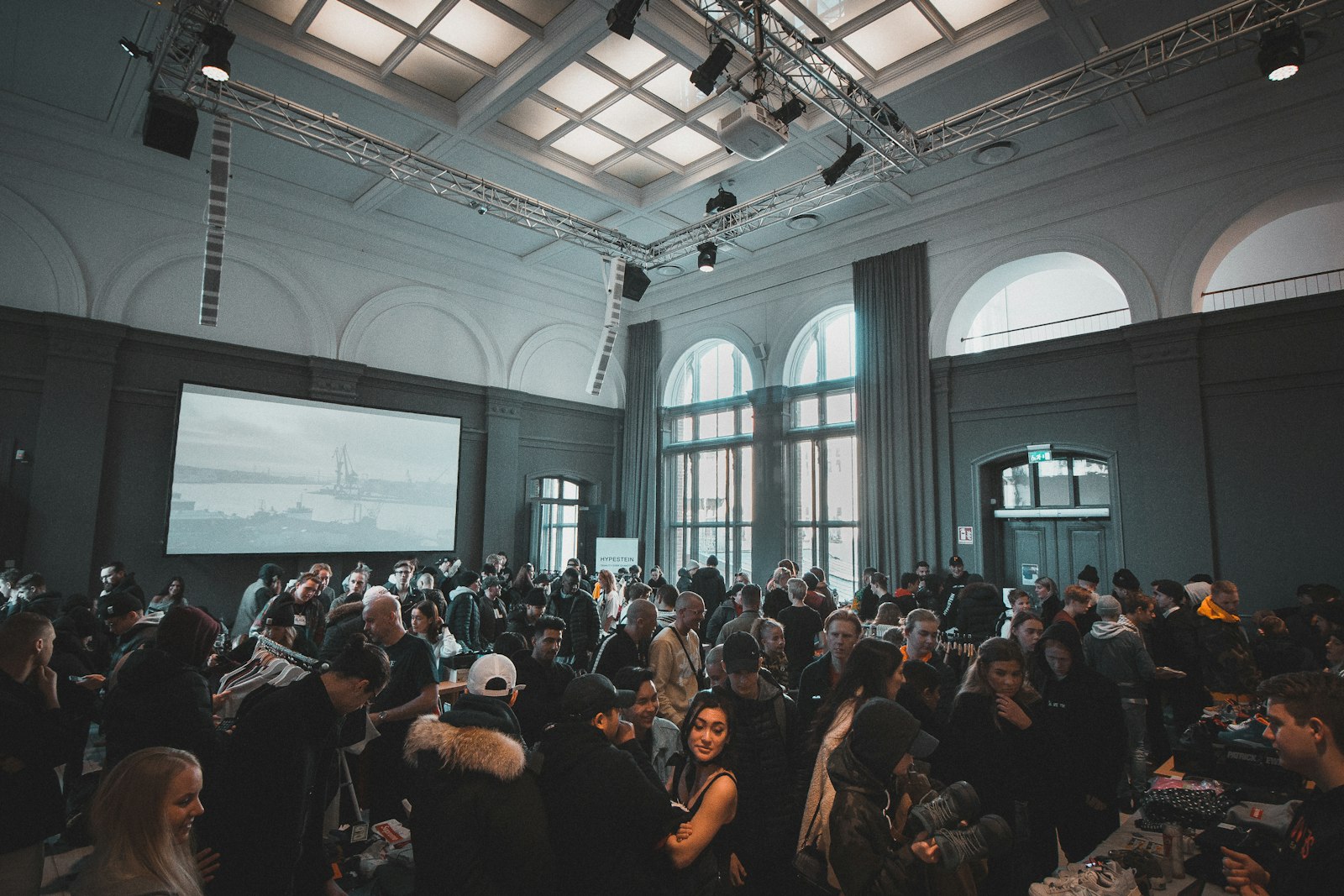 Standing crowd in a daylit hall with tall arched windows and a lighting truss
