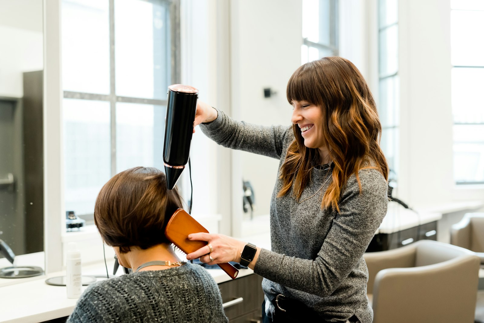 Smiling stylist blow-drying a client's bob with a round brush in a bright salon