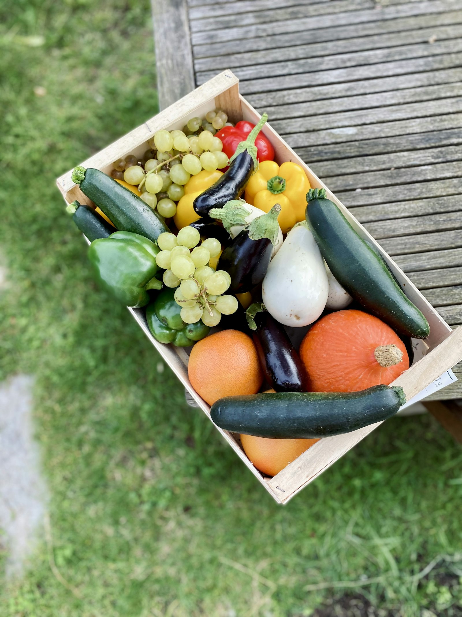 Wooden crate of peppers, courgettes, squash and grapes held over grass